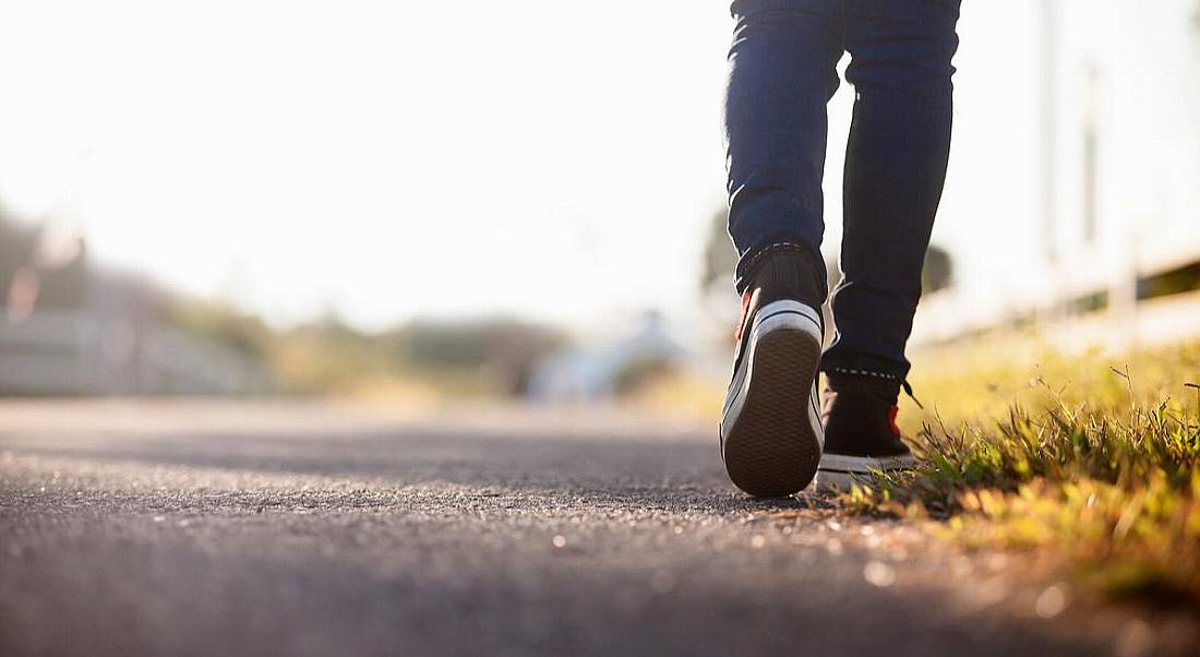 Close-up of the back of a person’s feet as they walk down an empty road. They are wearing dark blue jeans and black Converse.