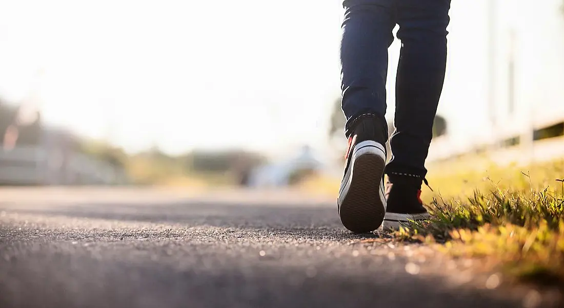 Close-up of the back of a person’s feet as they walk down an empty road. They are wearing dark blue jeans and black Converse.