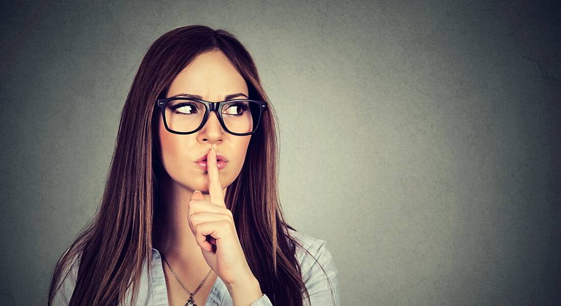 Brunette woman with finger on lips against a grey background, wondering who she can discreetly tell about her job search.