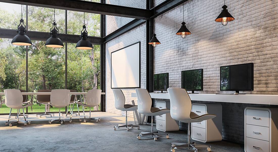 A view of a modern-looking office with wall-to-ceiling windows on one side, hanging lamps and an exposed brick accent wall.