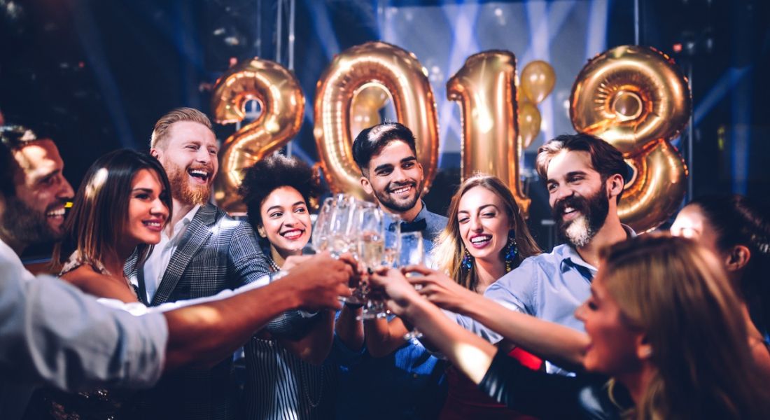 group of young adults laughing and smiling, toasting with glasses of champagne, in front of purple background with 2018 in gold balloons.