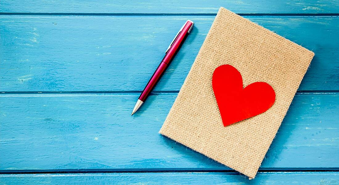 A fabric-covered notebook with a red heart on a blue wooden surface next to a metallic pink pen.