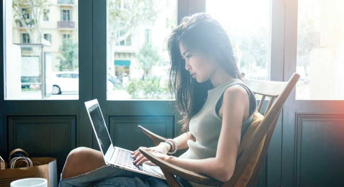 Young Asian woman sitting in a wooden rocking chair using her laptop. She is wearing a tank top and jeans