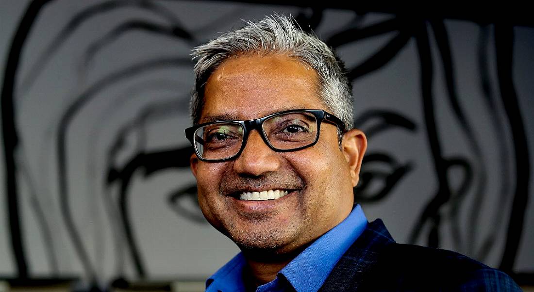 An Indian man with thick-framed glasses and silver stubble flashes a toothy smile at the camera against a grey background.