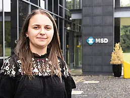 A brunette woman smiling at the camera wearing a black flowery blazer. The BMS office is blurred out behind her.