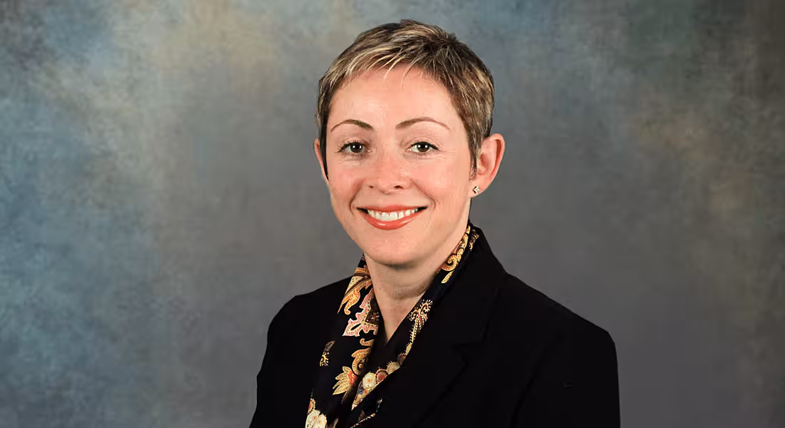 A woman with short, blonde hair and a black blazer smiling at the camera against a plain background.