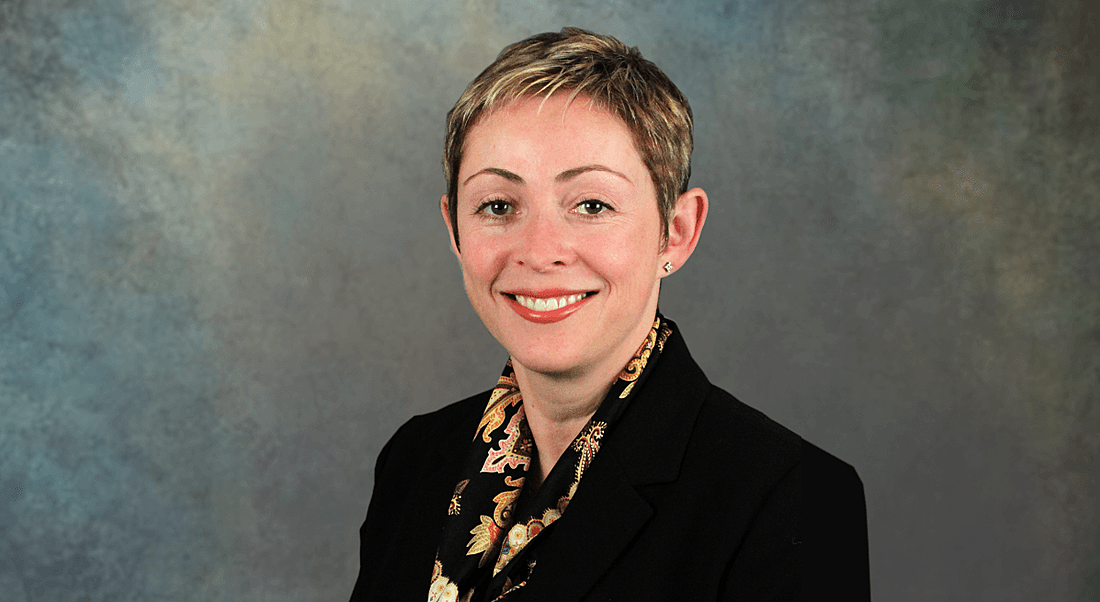 A woman with short, blonde hair and a black blazer smiling at the camera against a plain background.