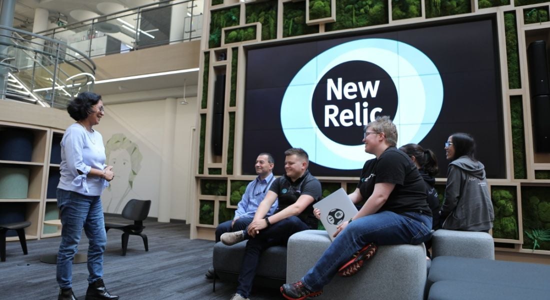 A diverse group of New Relic employees sitting together in the open plan officeon comfy stools facing one standing employee.