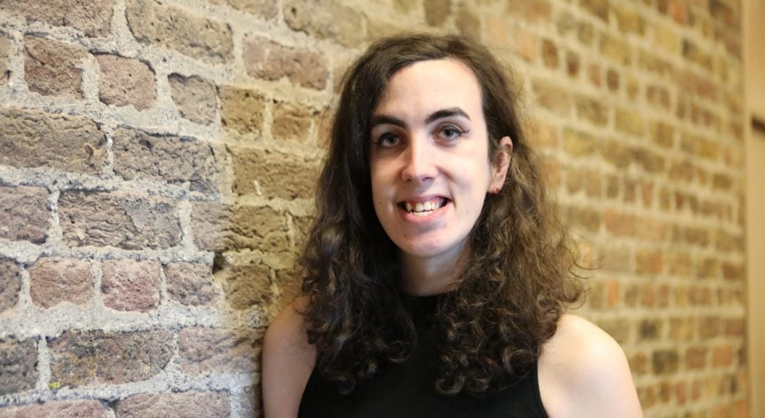 A curly-haired woman in a sleeveless black top smiles next to a brick wall.