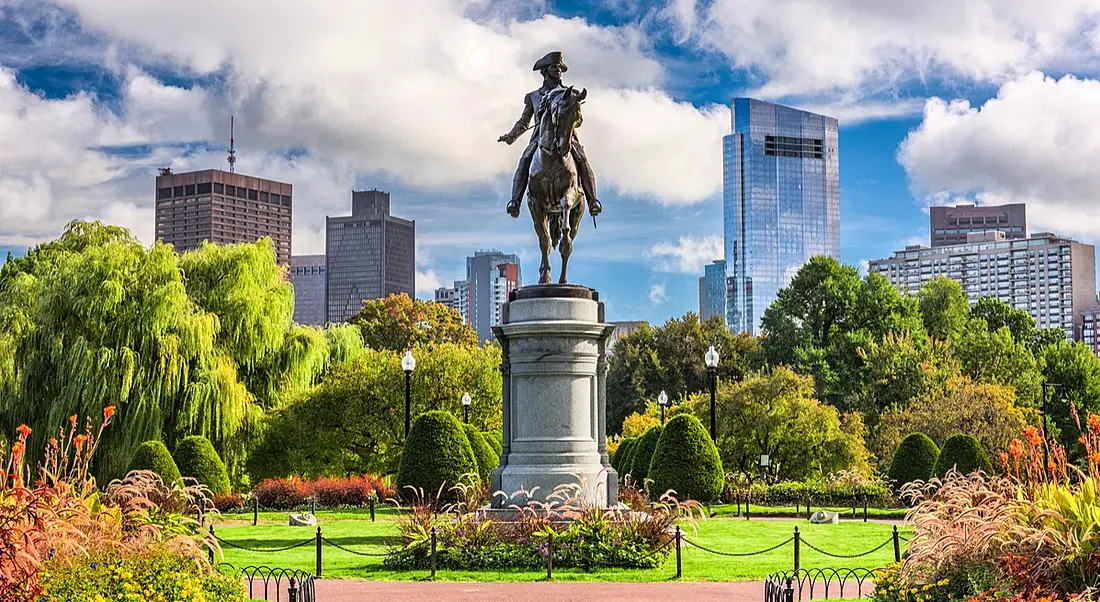 George Washington monument in the centre of a vibrant green park surrounded by trees with Boston skyscrapers behind it.