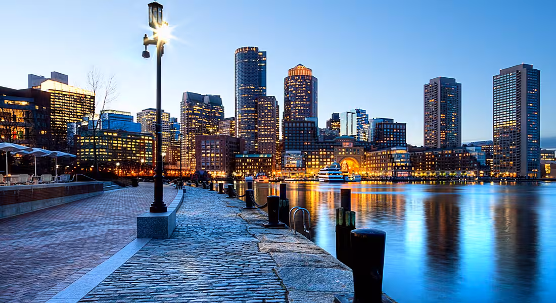 Low angle shot of Boston harbour and facing the lit-up, looming skyscrapers against a dusky sky.