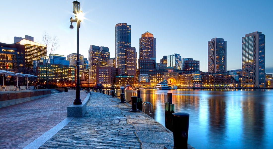 Low angle shot of Boston harbour and facing the lit-up, looming skyscrapers against a dusky sky.