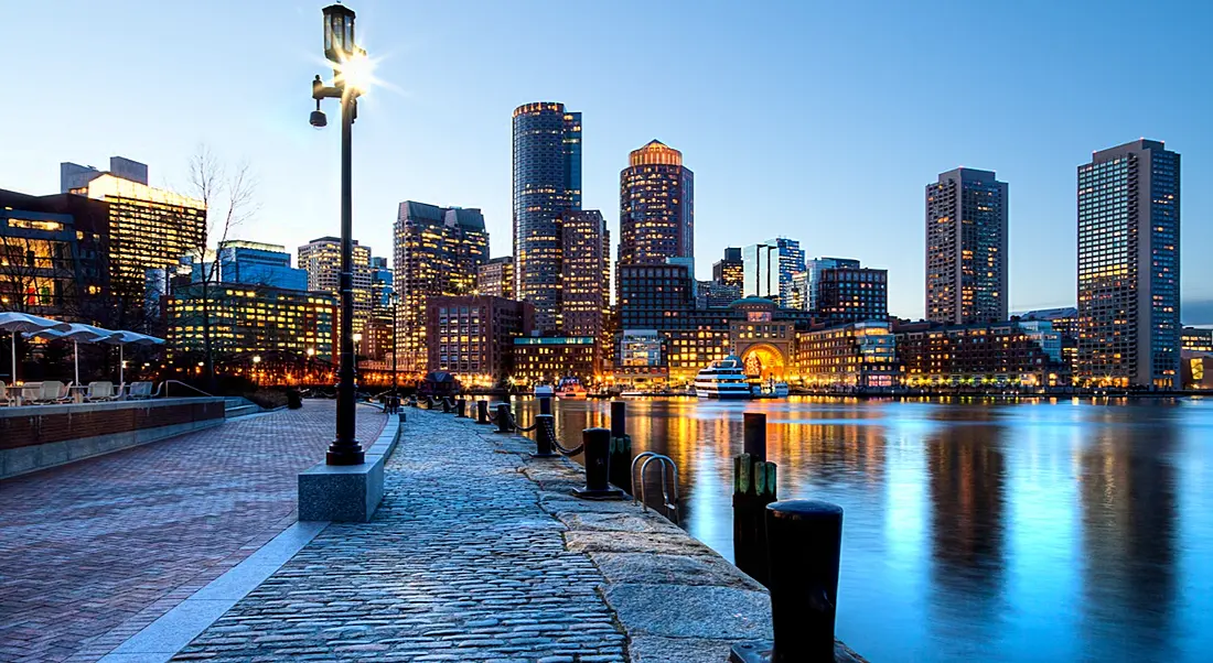 Low angle shot of Boston harbour and facing the lit-up, looming skyscrapers against a dusky sky.