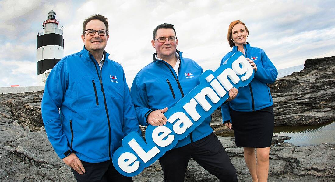 Three people in electric blue windbreakers holding a sign that says ‘eLearning’. On a rocky shore with a lighthouse visible behind them.