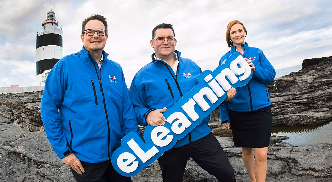 Three people in electric blue windbreakers holding a sign that says ‘eLearning’. On a rocky shore with a lighthouse visible behind them.