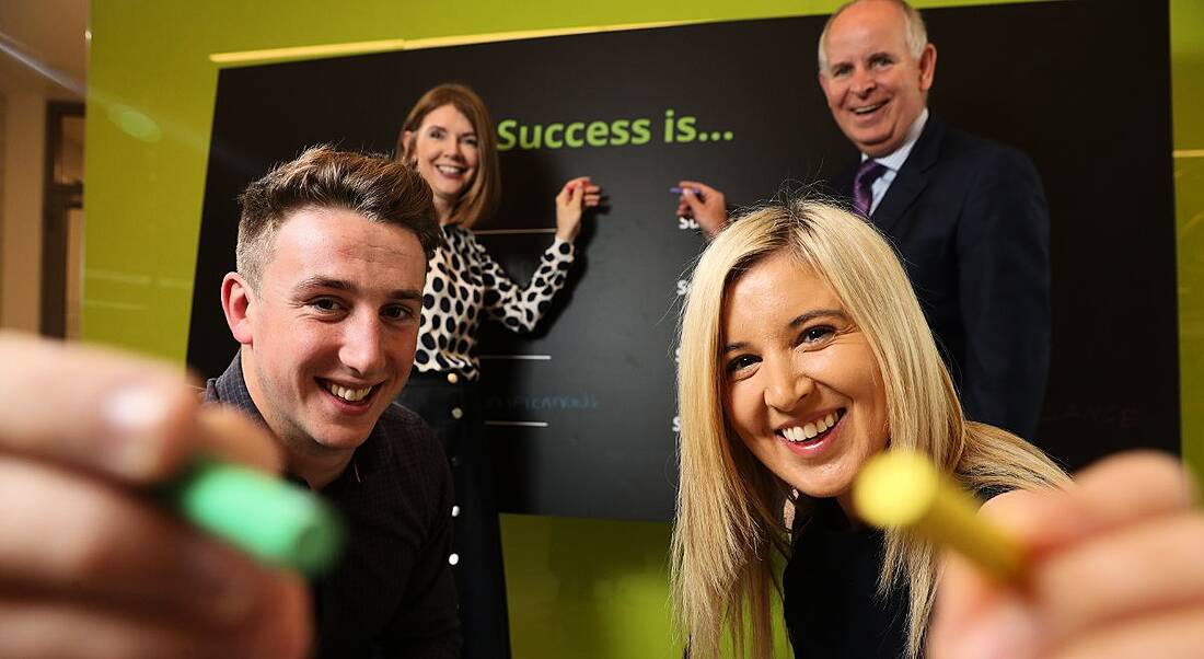 Two young graduates in foreground hold pieces of chalk while two older professionals point to a blackboard in the background.