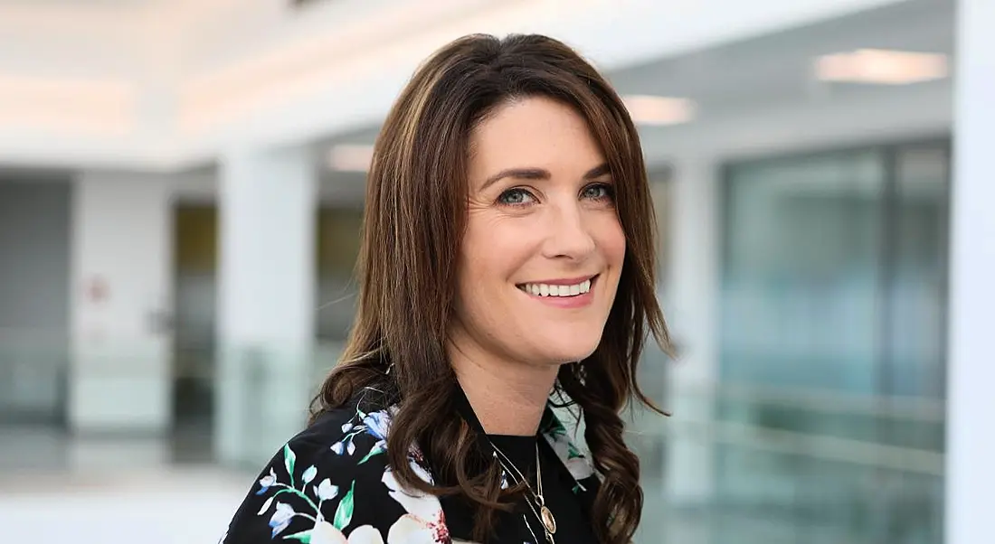 A brunette woman smiling at the camera wearing a black flowery blazer. The BMS office is blurred out behind her.