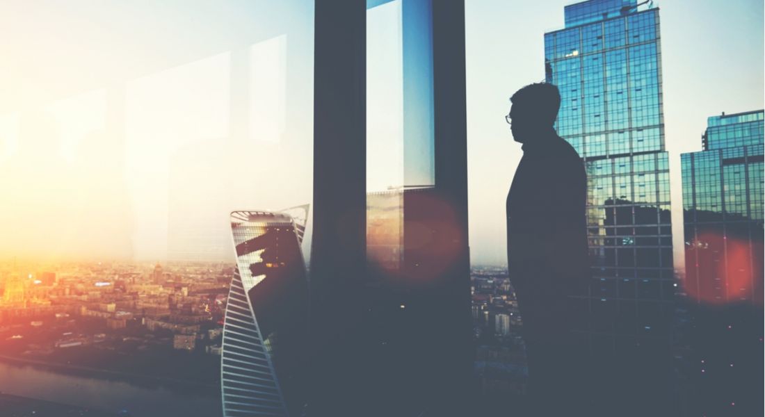 A silhouette of a man thinking about the future of work on a balcony looking out at a cityscape, with the sun rising.