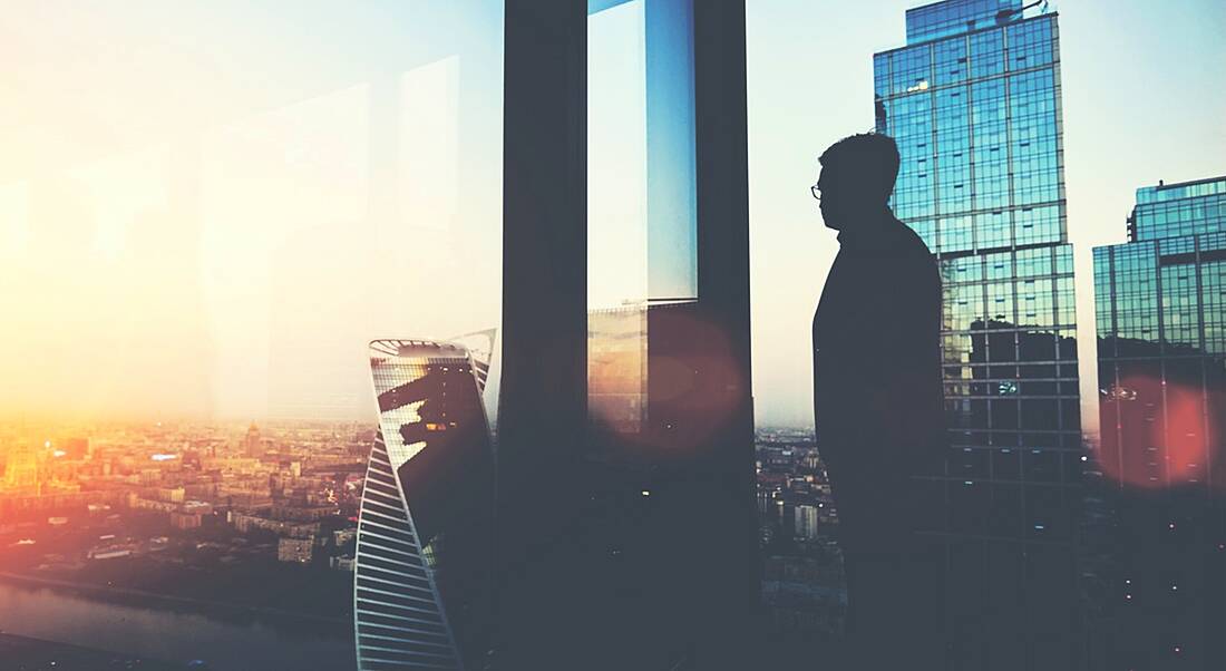 A silhouette of a man thinking about the future of work on a balcony looking out at a cityscape, with the sun rising.