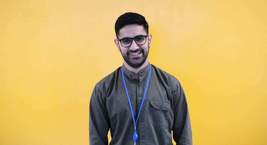 A smiling dark-haired man with a beard and glasses against a yellow background. He’s wearing a casual shirt and blue lanyard.