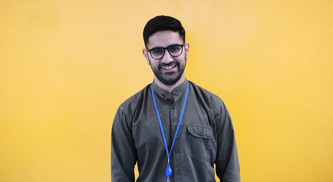 A smiling dark-haired man with a beard and glasses against a yellow background. He’s wearing a casual shirt and blue lanyard.