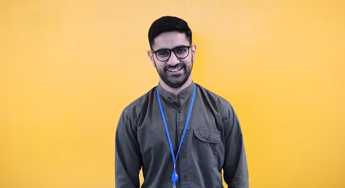 A smiling dark-haired man with a beard and glasses against a yellow background. He’s wearing a casual shirt and blue lanyard.