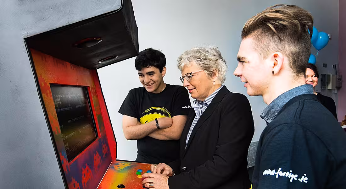 A woman and two teenage boys play on an arcade games console.