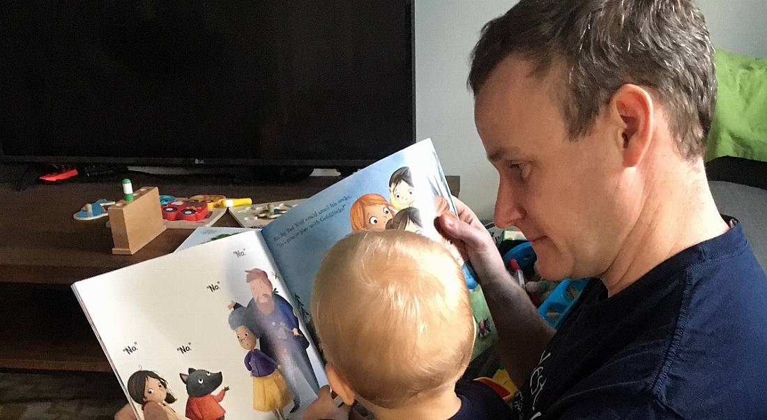 A man with short brown hair reading a children’s book to his toddler on paternity leave. The boy has his back to the camera.