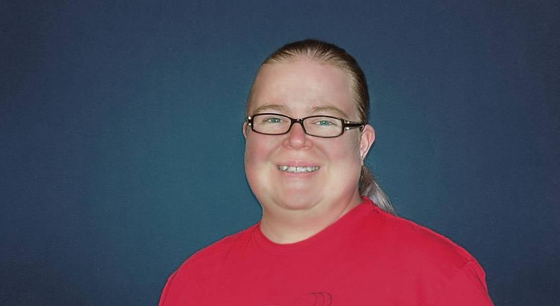 A woman with blonde hair tied back, wearing black glasses and a red t-shirt smiling at the camera against a navy background.