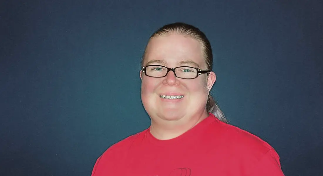 A woman with blonde hair tied back, wearing black glasses and a red t-shirt smiling at the camera against a navy background.