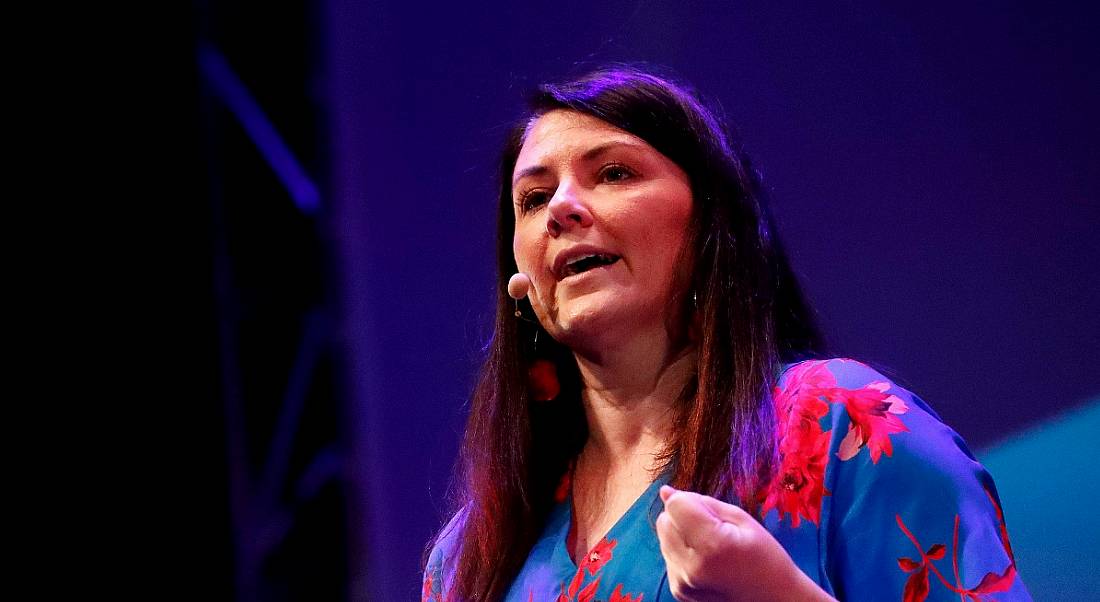 A close-up of a confident, dark-haired woman speaking at a conference about the future of work.