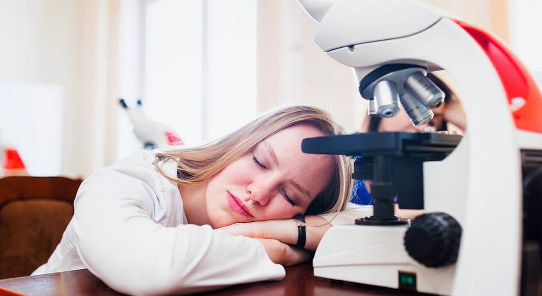 Blonde woman in a lab coat, napping on her desk in front of a microscope. She is stuck in a rut in her life sciences career.