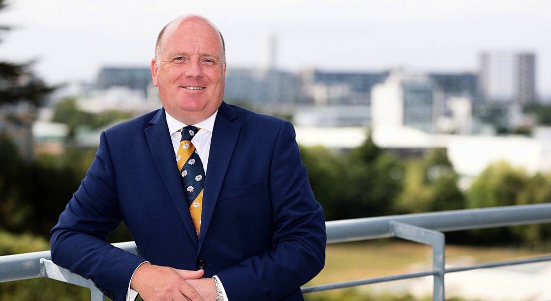 A jovial looking man in a navy suit leaning against a balcony railing.