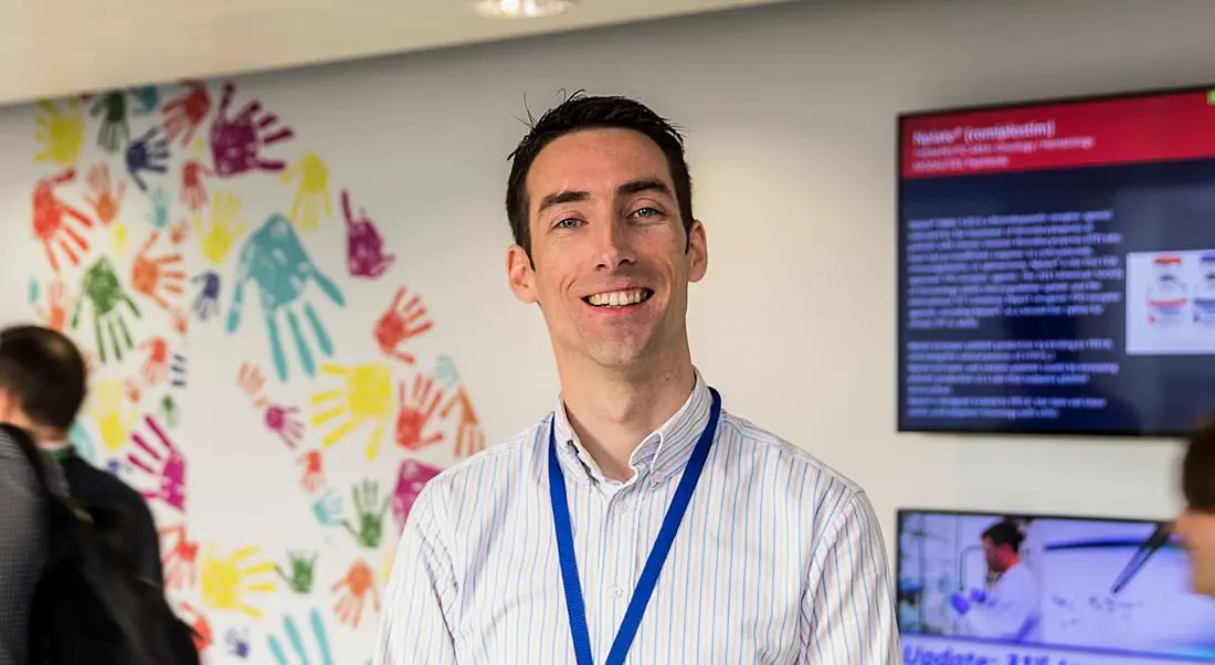 A young man in a powdery blue shirt grinning at the camera in Amgen.