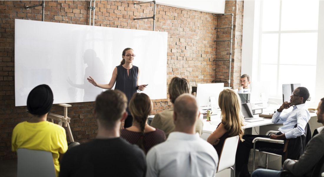 A woman standing in front of a seated crowd of professionals who are there for training. There’s a whiteboard behind her.