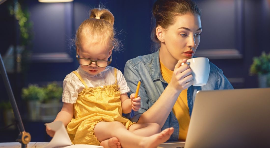 Woman holding coffee and staring at a laptop. Her daughter sits on the table beside her. This depicts working mothers.