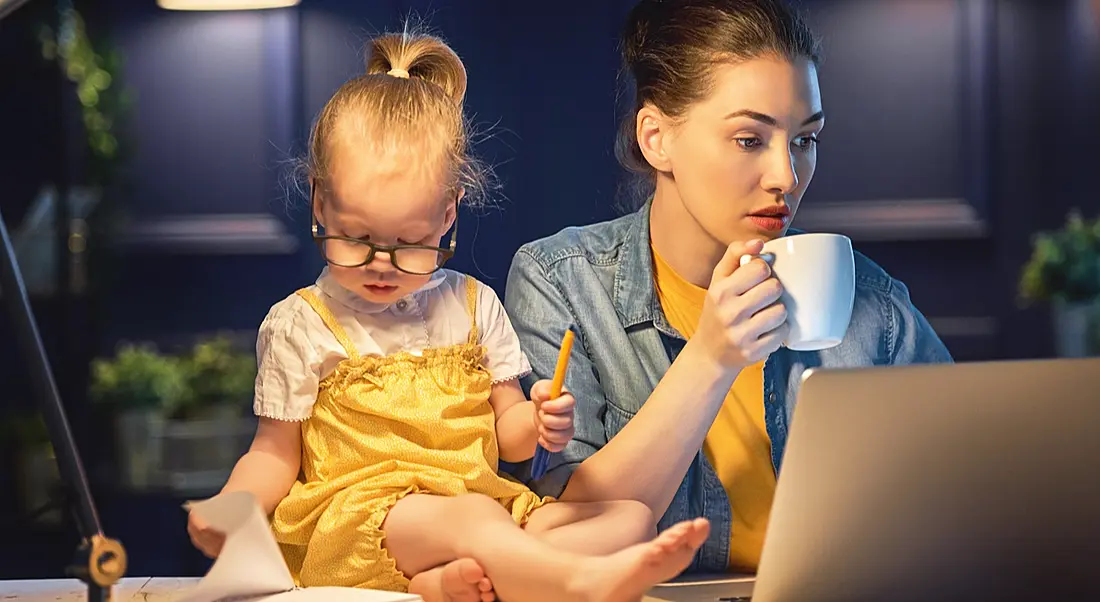 Woman holding coffee and staring at a laptop. Her daughter sits on the table beside her. This depicts working mothers.