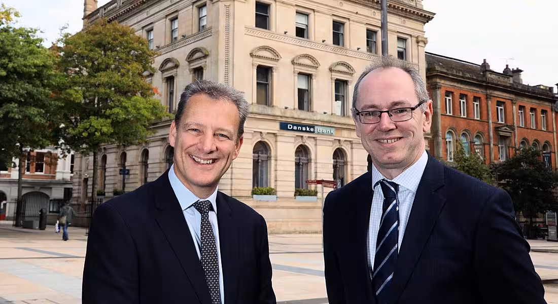 Two middle-aged men in suits looking directly into the camera and smiling widely against the backdrop of Danske Bank.
