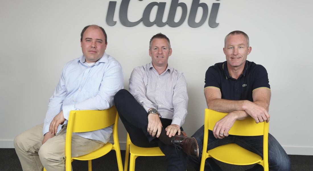 Three middle aged men sitting on bright yellow chairs underneath a logo emblazoned iCabbi in elegant italic font.