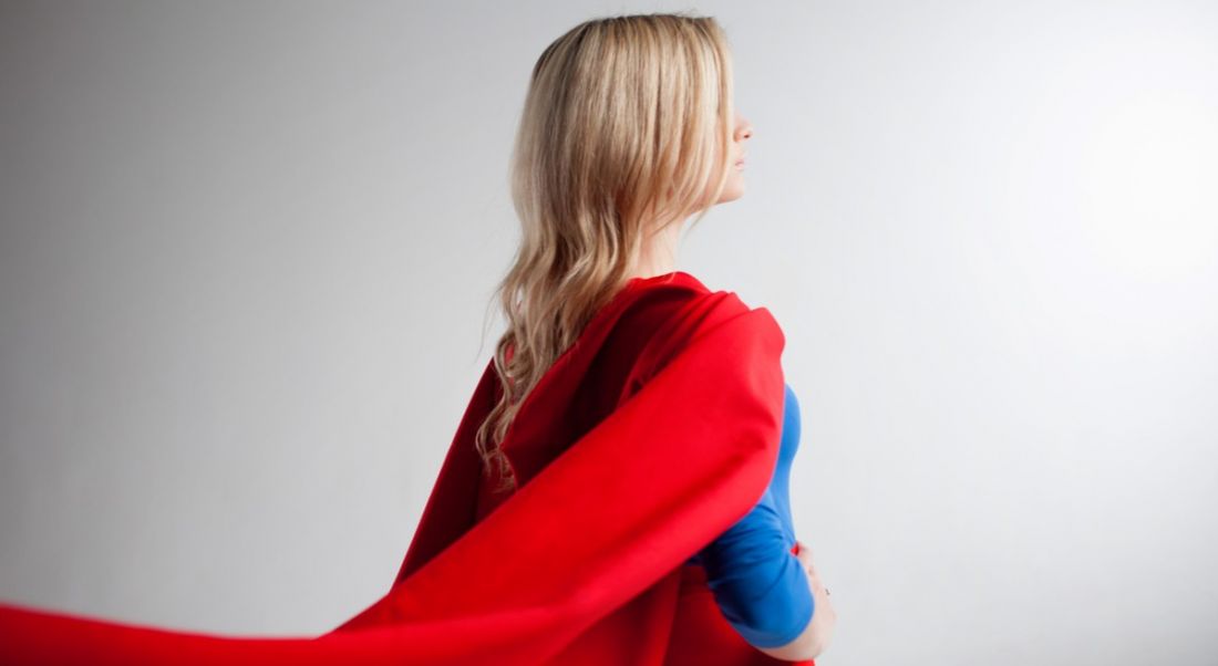 A blonde woman in a blue top, red skirt and red cape stands with her back to the viewer.