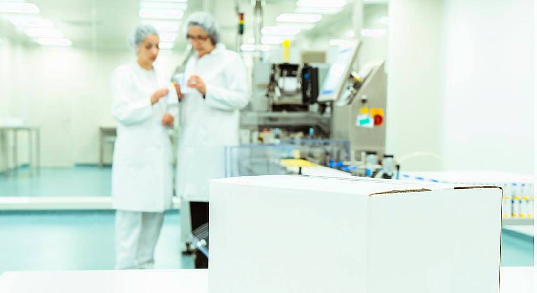 white box on table in forefront with two pharma workers in blurry background wearing lab coats and hair nets.