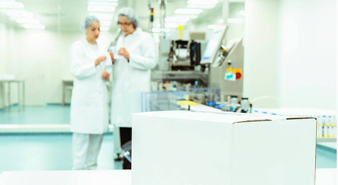 white box on table in forefront with two pharma workers in blurry background wearing lab coats and hair nets.