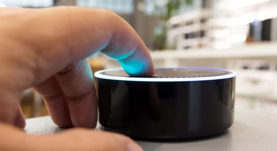 Close-up of a hand reaching for a button on an Amazon Echo device which is lit up by a ring of blue light.