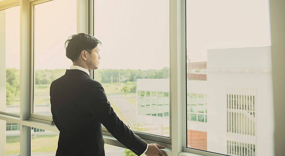 A young man in a dark business suit staring pensively out a floor-to-wall window thinking about the future of work.