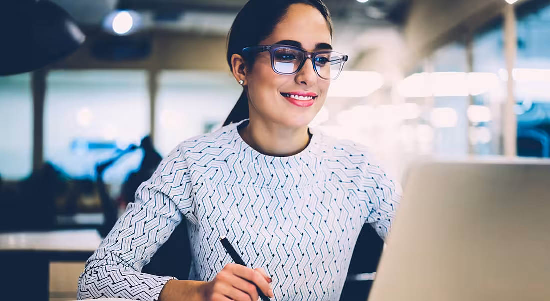 Young woman with glasses smiling at a laptop. She’s writing down the future of work influencers she needs to follow.