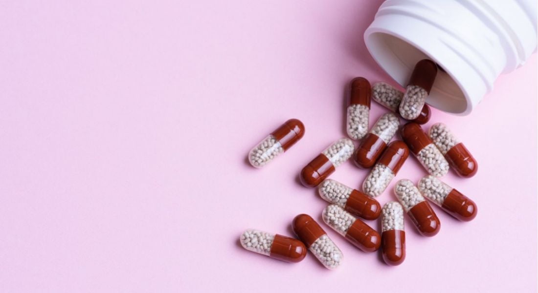Red and white pill capsules spilling out of a white bottle on a baby pink background