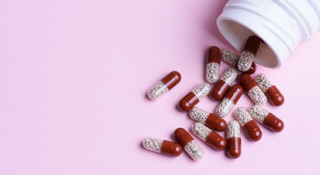 Red and white pill capsules spilling out of a white bottle on a baby pink background