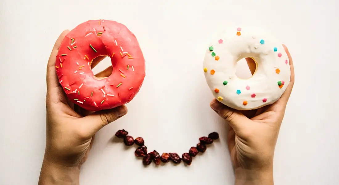 Two hands holding iced donuts over a row of coffee beans, making the appearance of a smiley face.