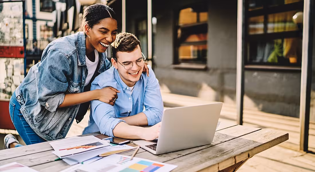 two friends, a male and a female, huddled in front of a laptop and smiling.