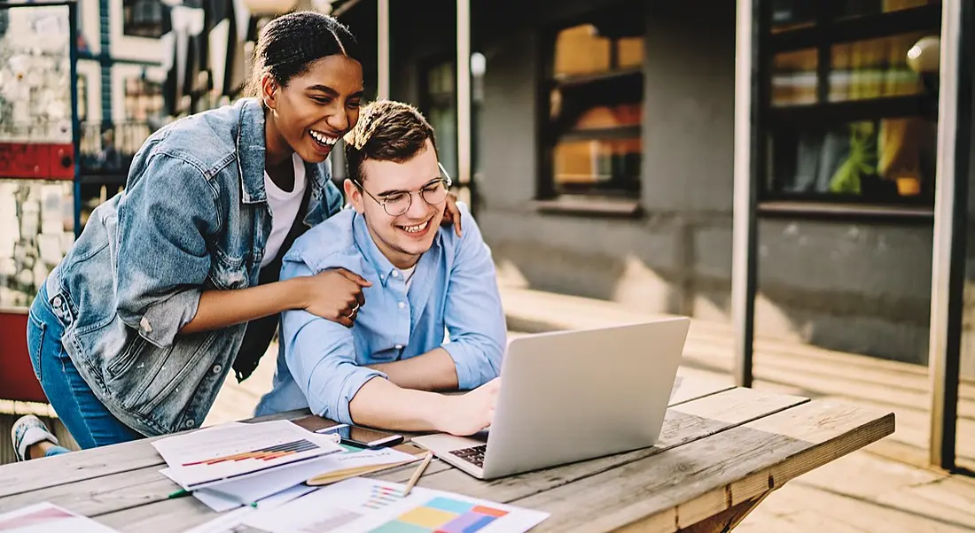 two friends, a male and a female, huddled in front of a laptop and smiling.